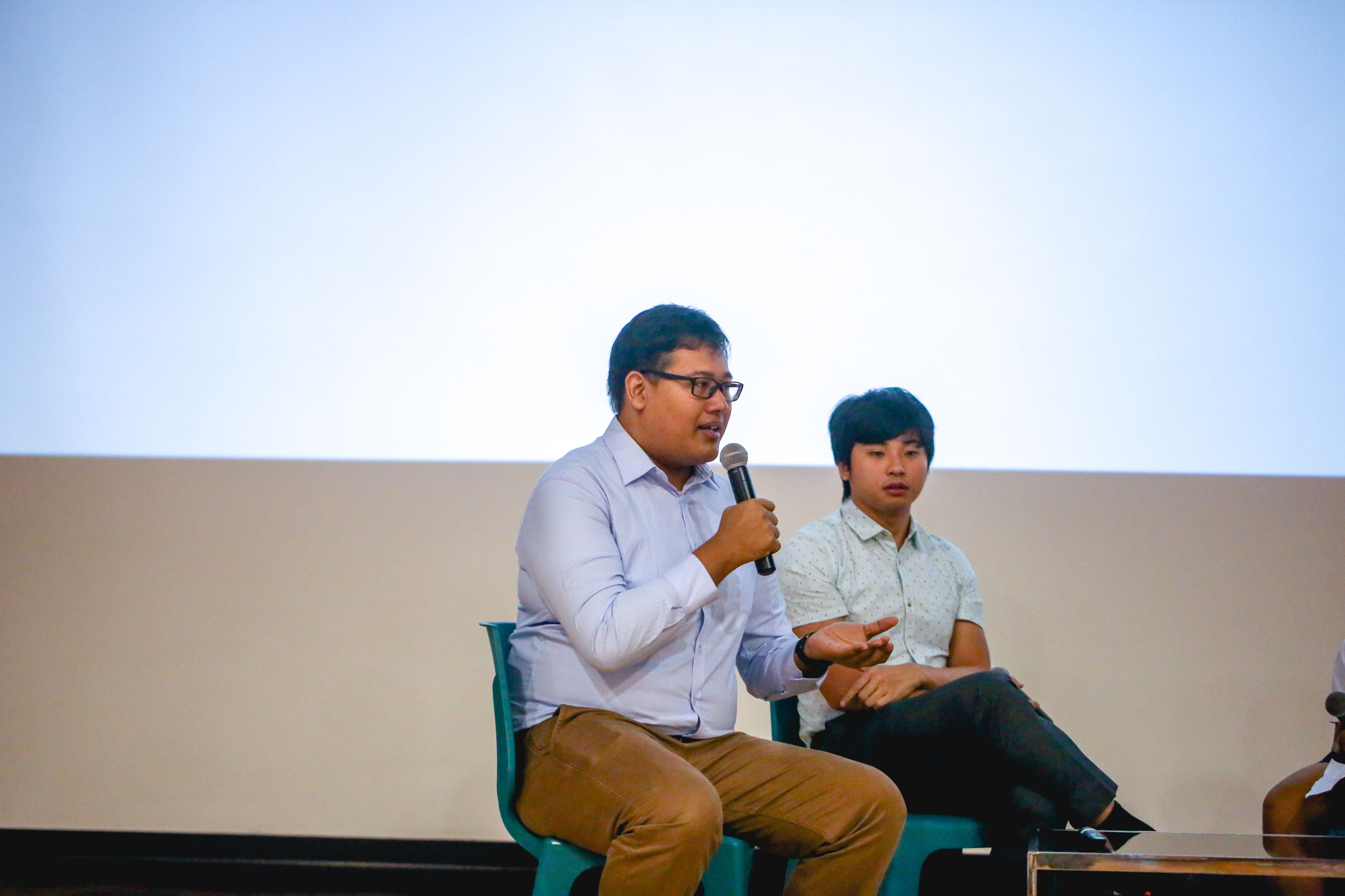Man with glasses speaking into a microphone while seated, with another young man listening beside him on stage.